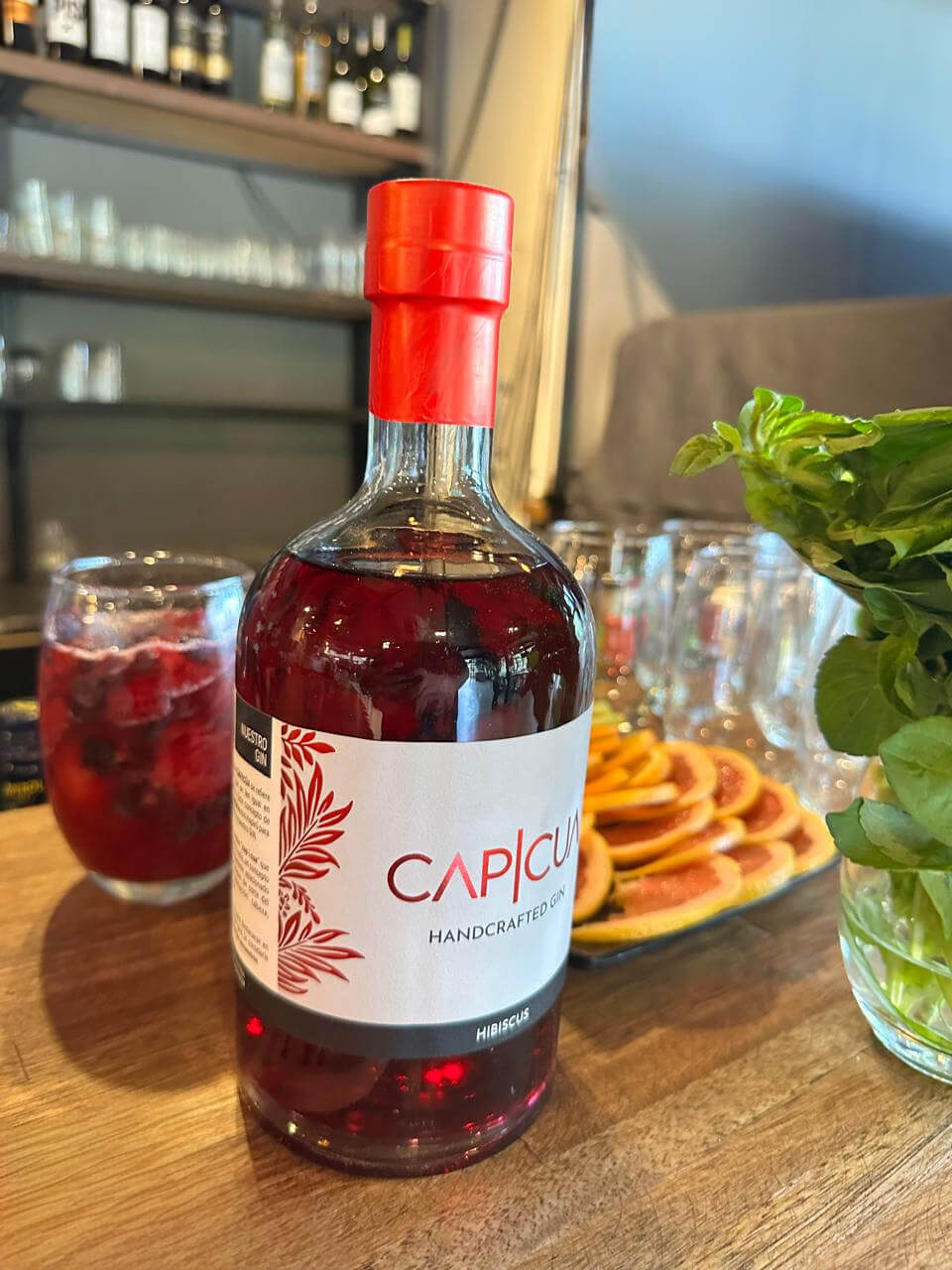 Botella CAPICÚA Hibiscus presentada en una barra junto a cítricos y un cóctel